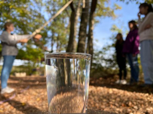 Ein Glas Wasser im Vordergrund, während Menschen im Hintergrund stehend beobachten.